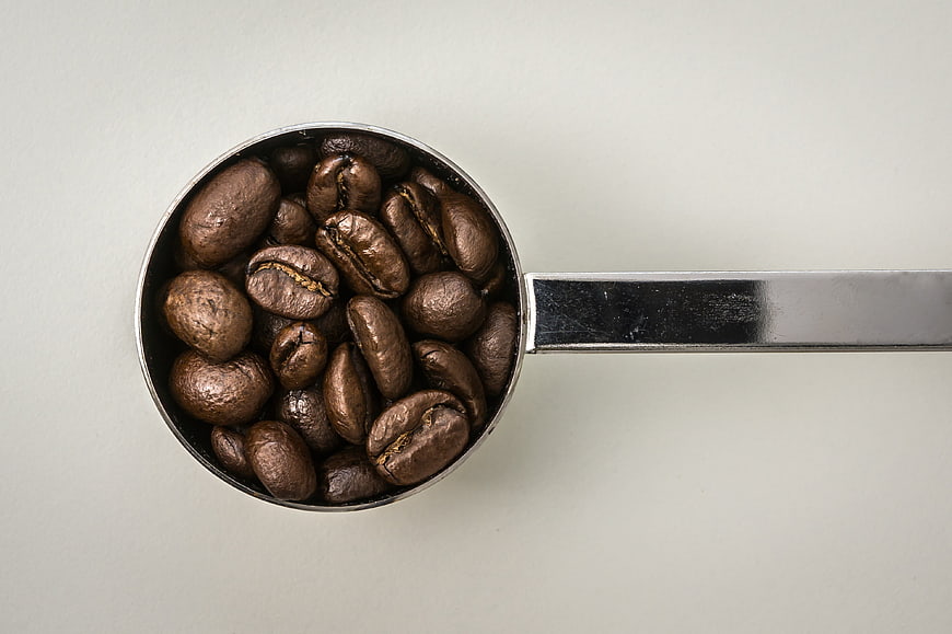Roasted coffee beans in a stainless steel measuring spoon on a light gray background