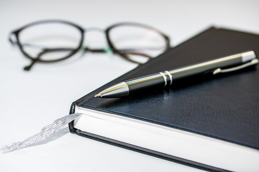 Black leather notebook with silver pen and reading glasses on white desk