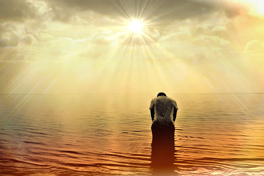 Man kneeling in ocean water praying under divine sun rays