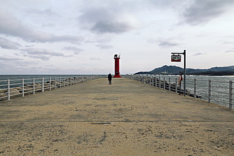 Lone figure walking on pier toward red lighthouse under cloudy sky