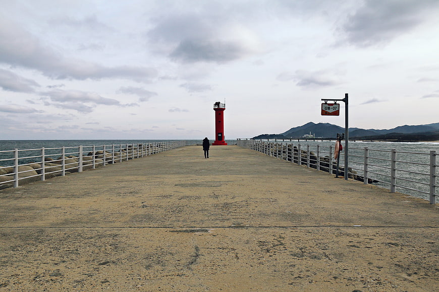 Lone figure walking on pier toward red lighthouse under cloudy sky