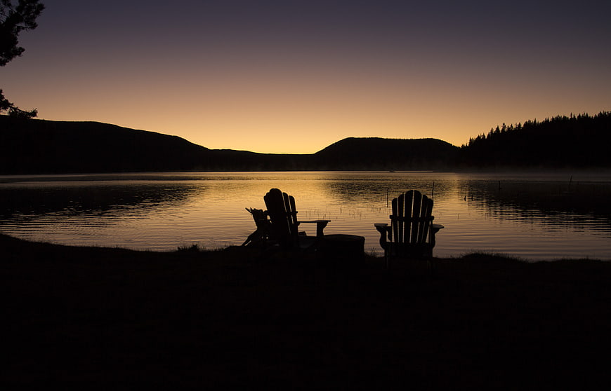 Silhouetted Adirondack Chairs by Lake at Sunset