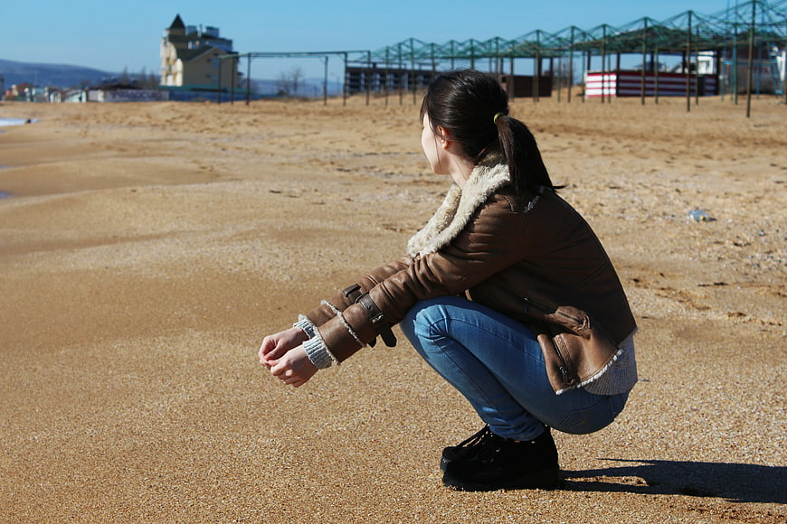 Woman Crouching on Sandy Beach in Winter Jacket