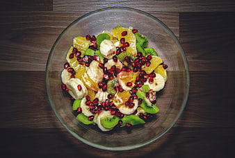Overhead view of fresh fruit salad with kiwi, banana, orange, and pomegranate in a glass bowl on a wooden table.