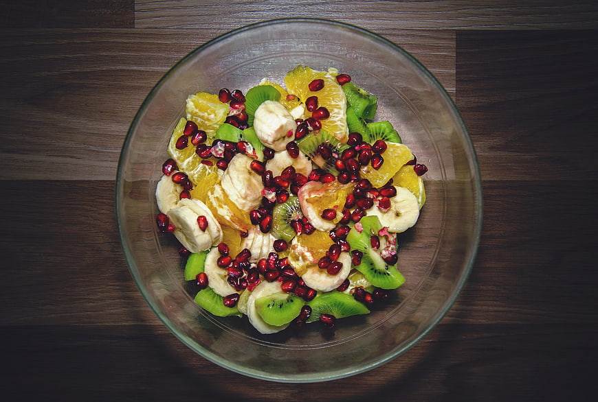 Overhead view of fresh fruit salad with kiwi, banana, orange, and pomegranate in a glass bowl on a wooden table.