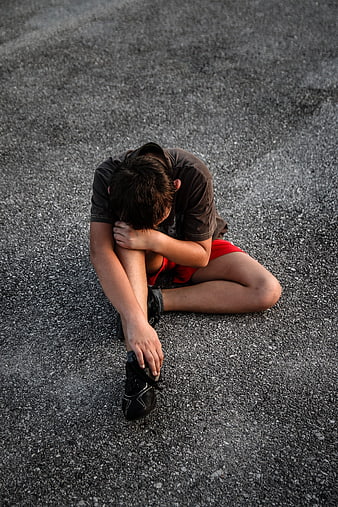 Sad boy sitting on asphalt holding his leg in pain