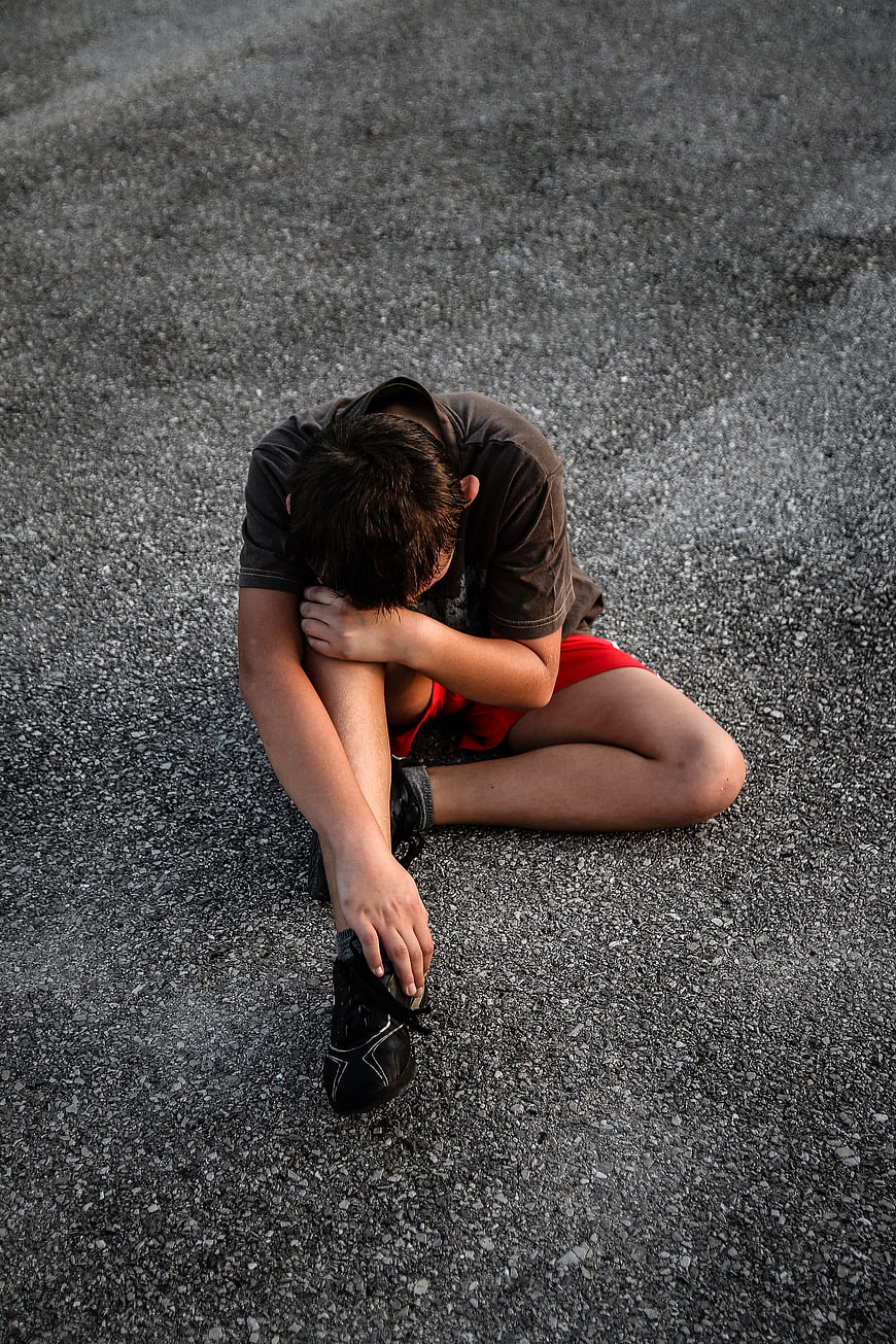 Sad boy sitting on asphalt holding his leg in pain