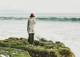 Woman in Hat Standing on Mossy Rocks by Ocean