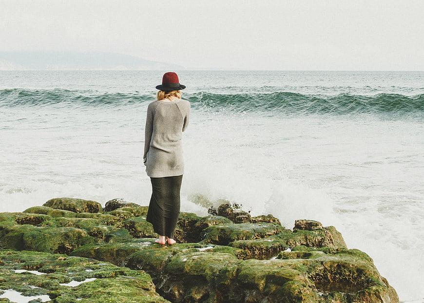 Woman in Hat Standing on Mossy Rocks by Ocean