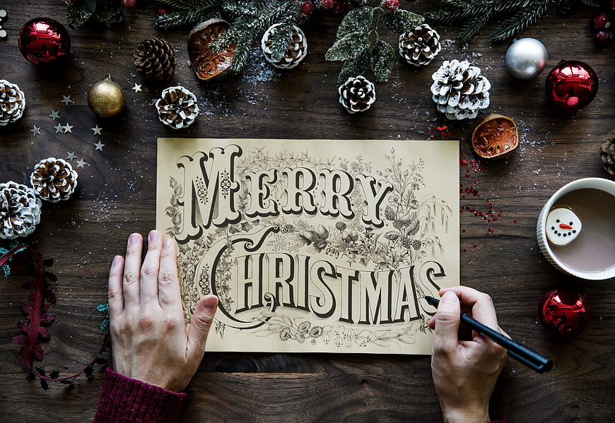 Hands drawing Merry Christmas lettering on paper with festive decorations