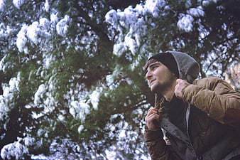 Young man in winter jacket looking up at snowy pine tree