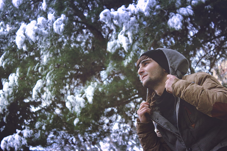 Young man in winter jacket looking up at snowy pine tree