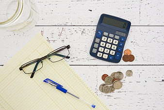 Top view of calculator, coins, glasses, and notebook on white wood