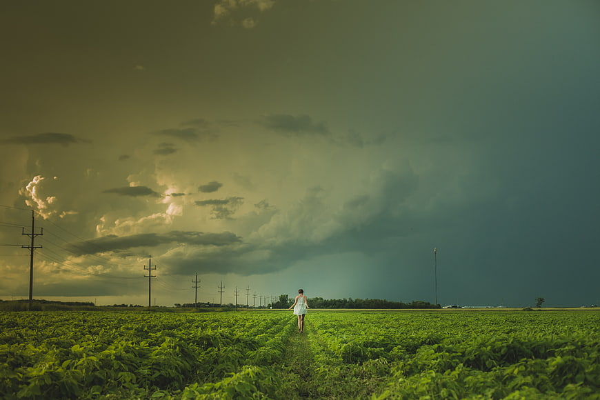 Woman in White Dress Walking Through Green Field Under Stormy Sky