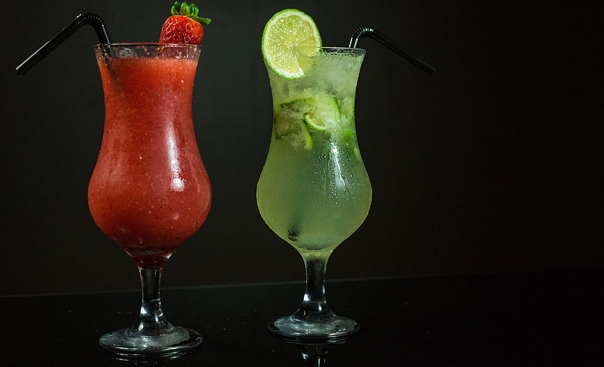 Two colorful cocktails in hurricane glasses with fruit garnish