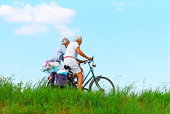 Elderly couple cycling together on a grassy hill under a bright blue sky