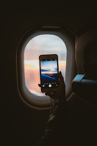 Hand holding smartphone taking photo of sunset through airplane window