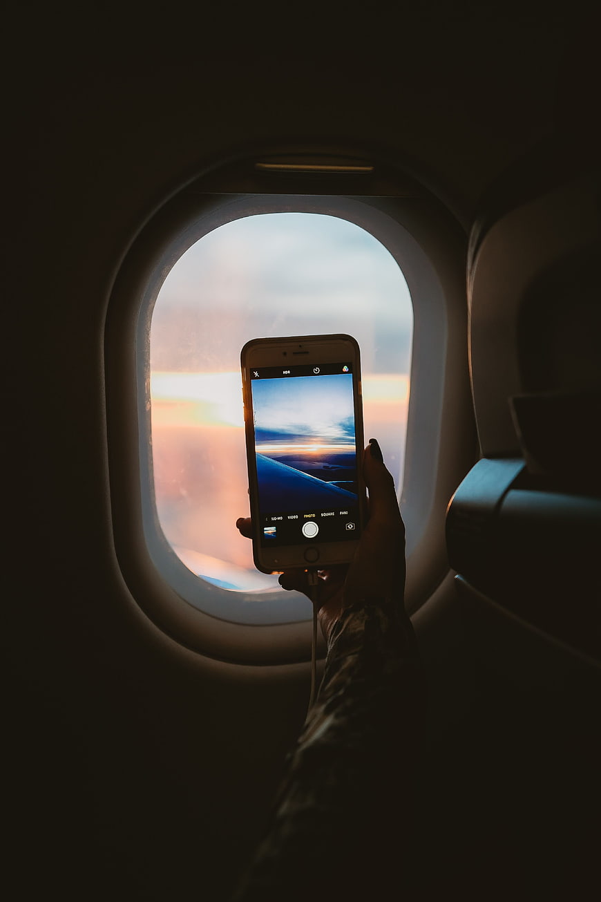 Hand holding smartphone taking photo of sunset through airplane window