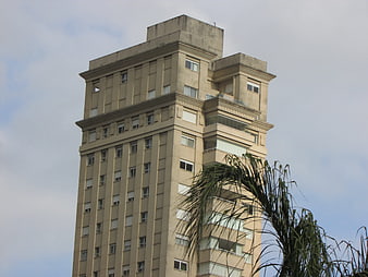 Tall beige apartment building with palm tree against cloudy sky