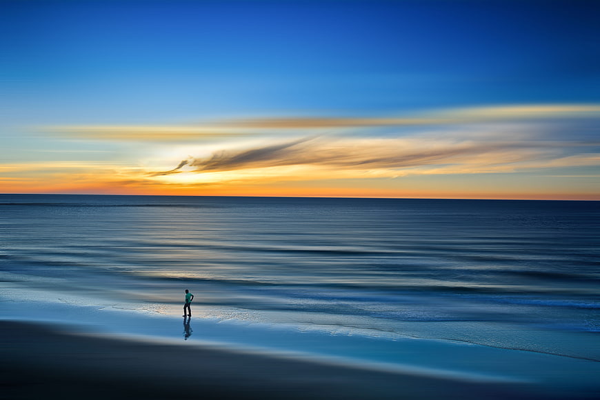 Solitary figure walking on a calm beach at sunset with long exposure water