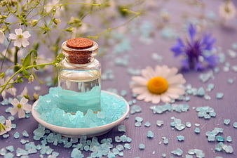 Glass essential oil bottle with blue sea salt and flowers on purple background