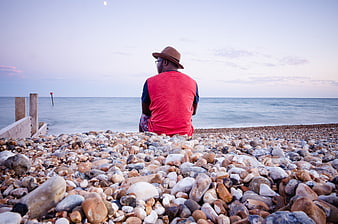 Man in Red Shirt Sitting on Rocky Beach at Sunset