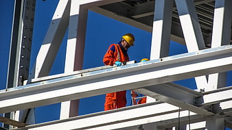 Construction worker in orange uniform and yellow hard hat on steel beams