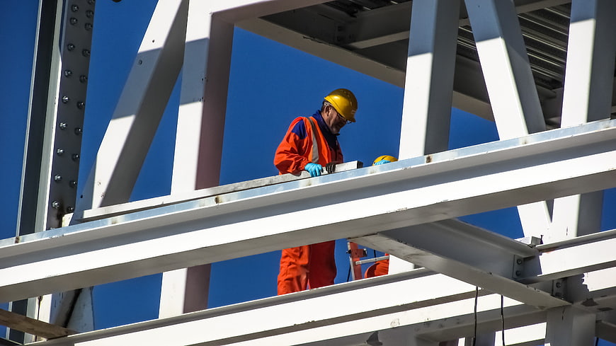Construction worker in orange uniform and yellow hard hat on steel beams
