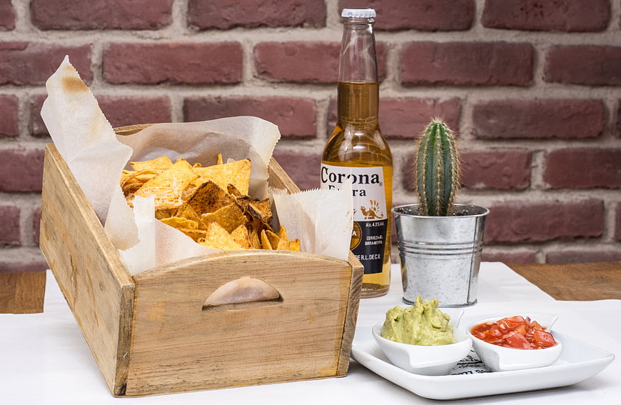 Wooden crate of nachos with Corona beer, salsa, and guacamole on a white table