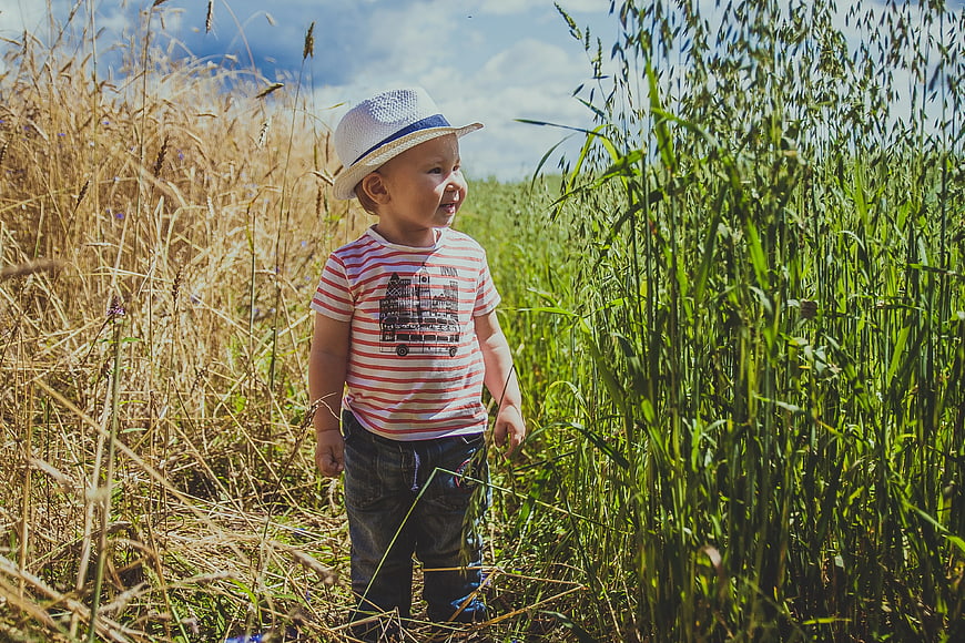 Toddler Boy in Straw Hat Standing in Tall Grass Field
