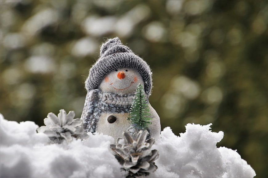 Cute Knitted Snowman Figurine in Snow with Pinecones and Bokeh Background