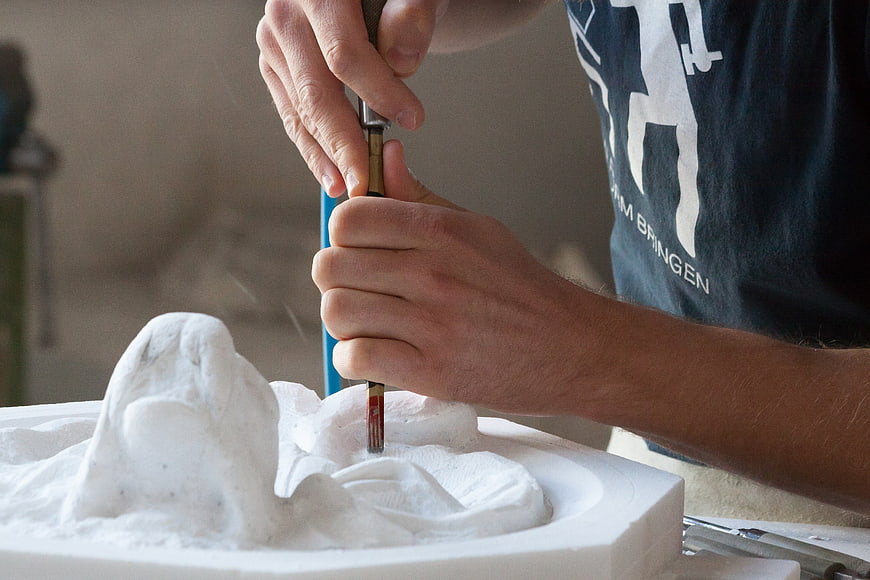 Close-up of hands using a pneumatic chisel to carve a white stone sculpture