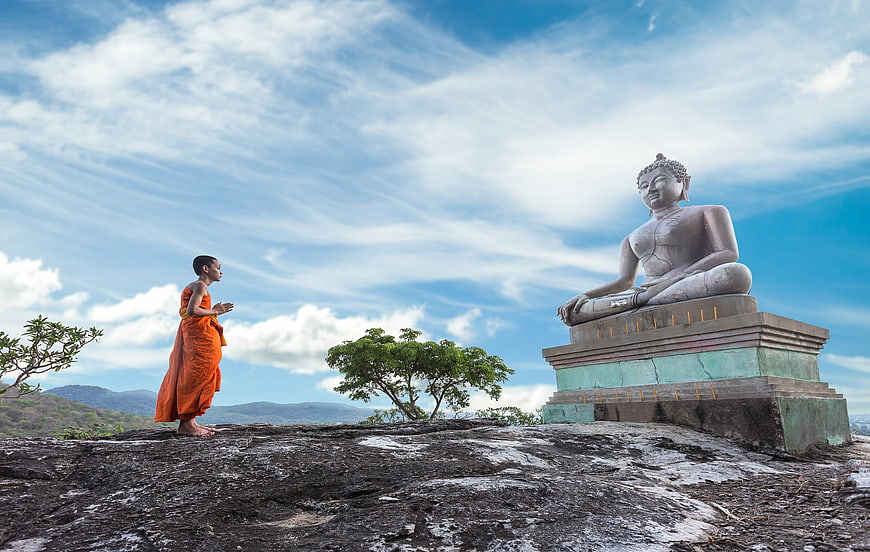 Young Buddhist Monk Praying Before Large Buddha Statue on Hilltop