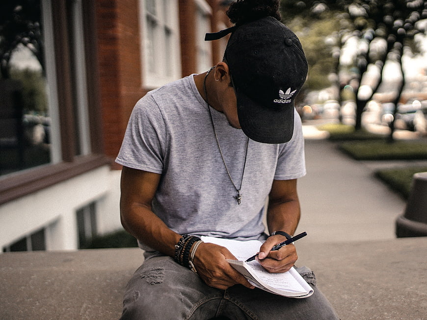 Man Writing in Notebook Outdoors Wearing Black Cap