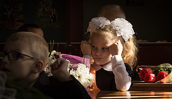 Young girl with white bows resting chin on hands in classroom