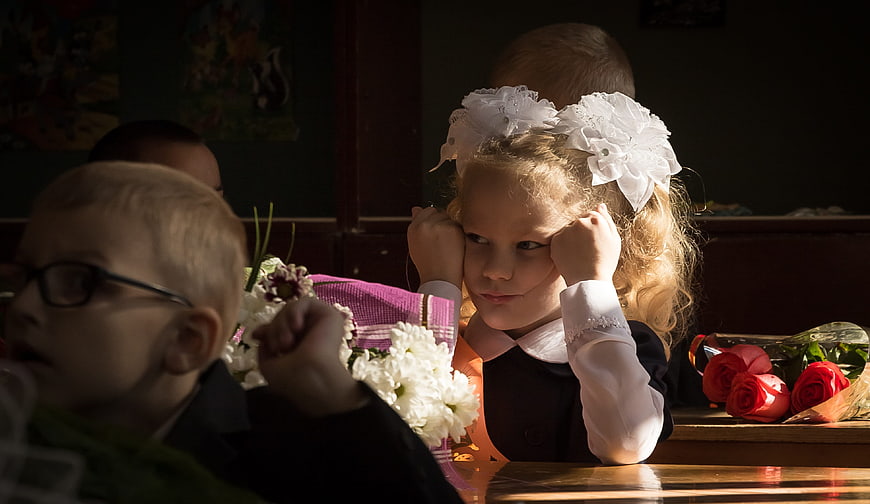 Young girl with white bows resting chin on hands in classroom