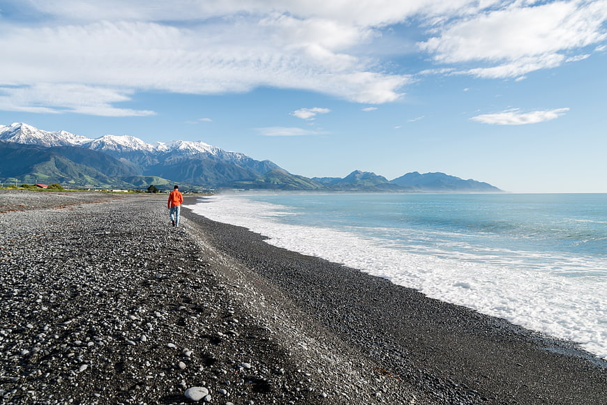 Man Walking on Pebble Beach with Snowy Mountains