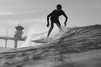 Surfer riding wave near pier in black and white