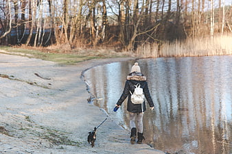 Woman walking small dog on sandy riverbank in winter