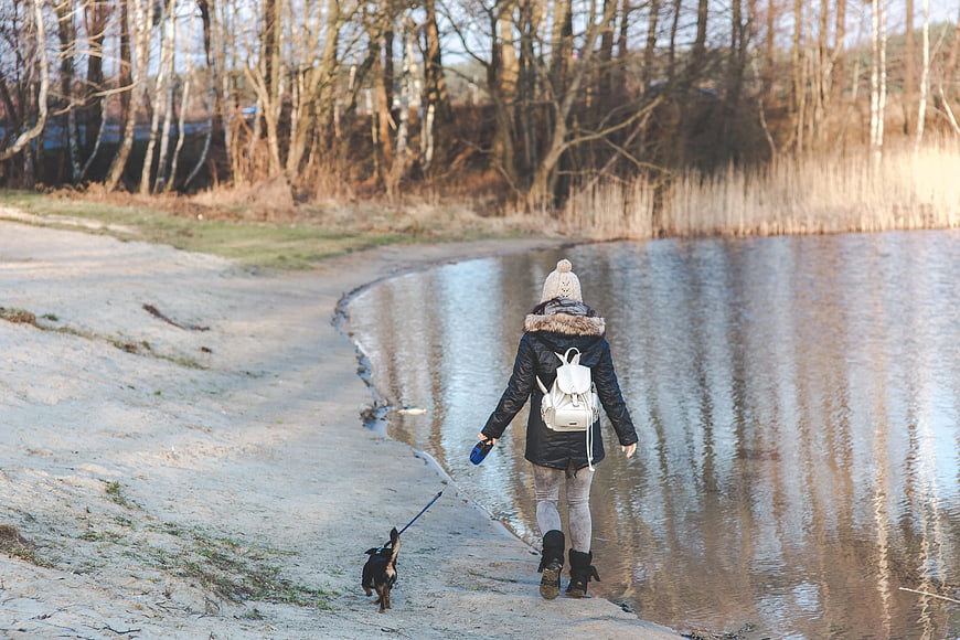 Woman walking small dog on sandy riverbank in winter