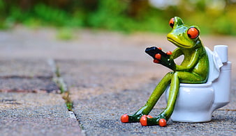 Cute Green Frog Figurine Sitting on Miniature Toilet with Smartphone