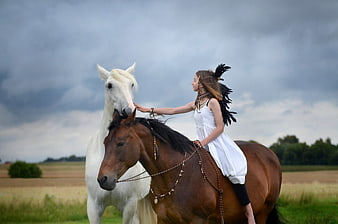 Girl in white dress riding brown horse touching white horse in field