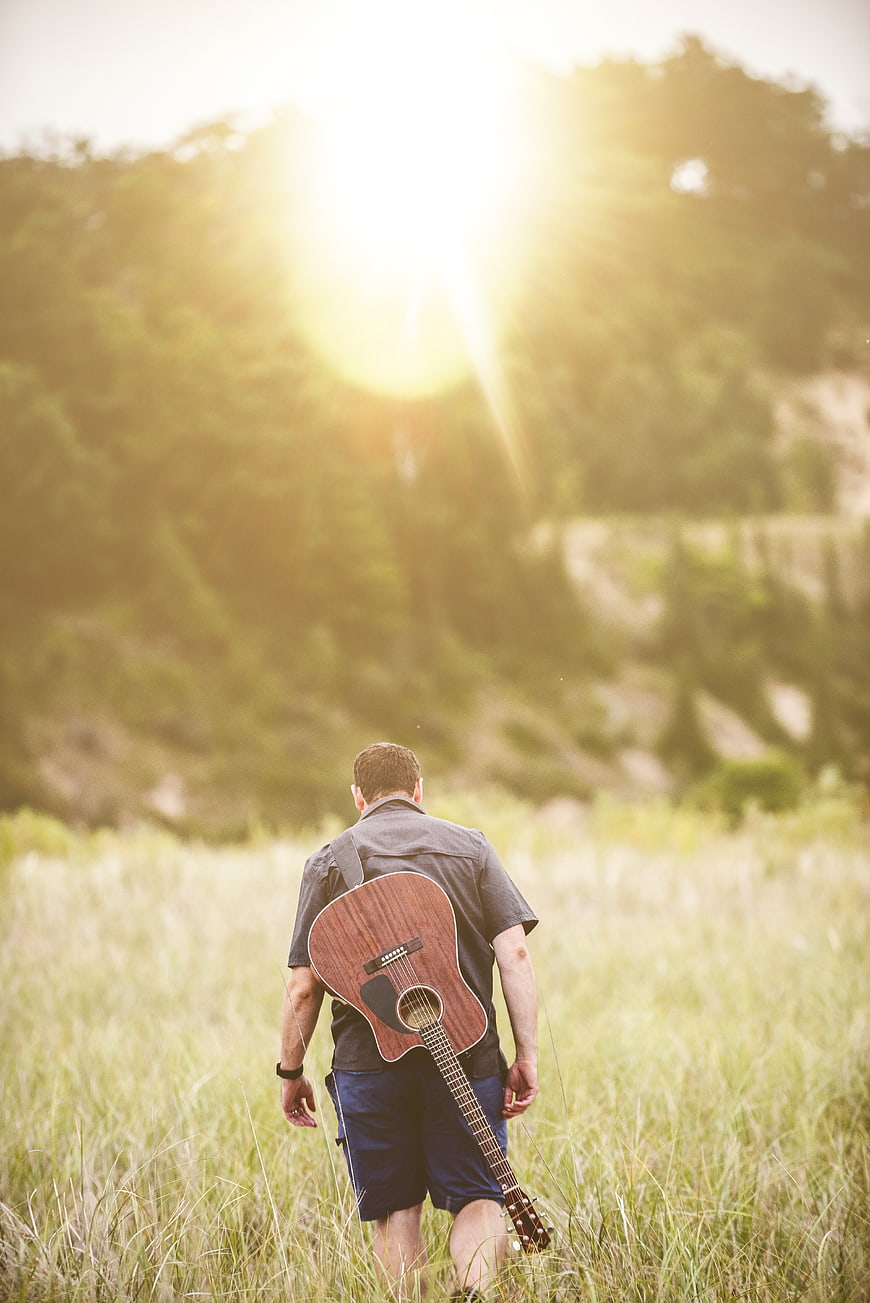 Man with Guitar Walking in Grassy Field at Sunset
