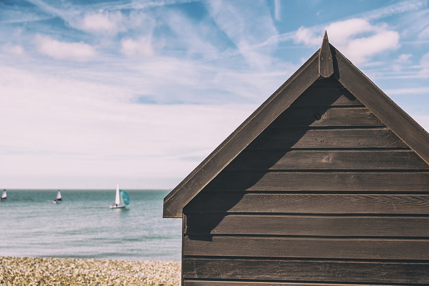 Dark Wooden Beach Hut on Pebble Shore with Sailboats