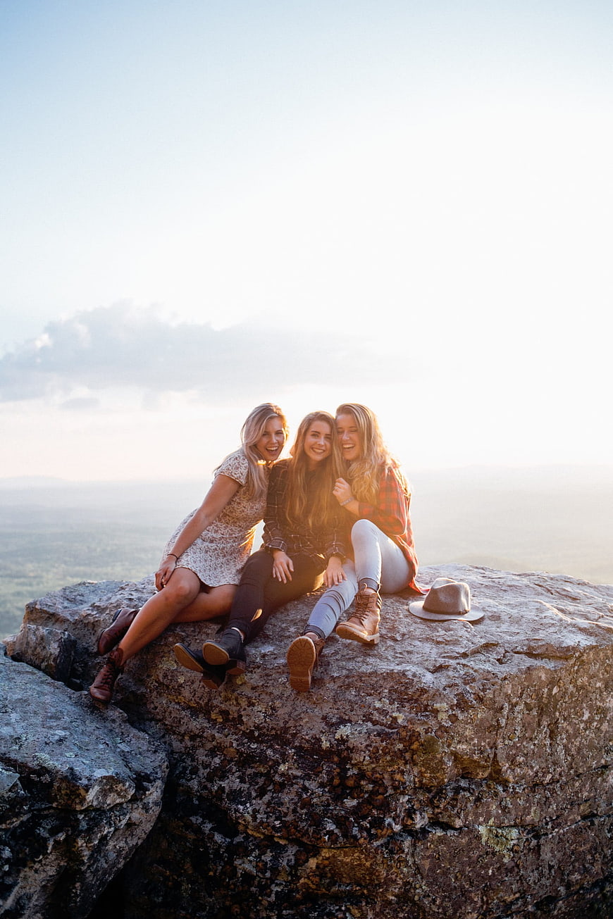 Three smiling female friends sitting on a rocky cliff edge at sunset