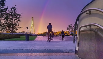 Cyclist watching rainbow over modern bridge at sunset