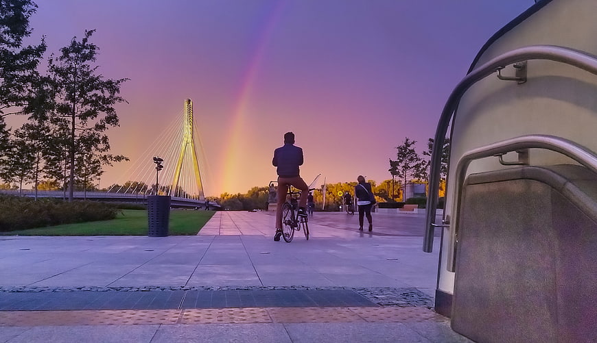 Cyclist watching rainbow over modern bridge at sunset
