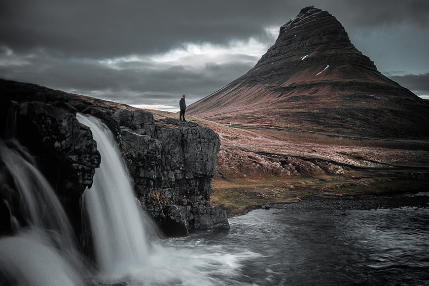 Lone hiker standing on cliff near waterfall with Kirkjufell mountain