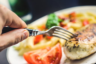 Close-up of hand holding fork over grilled chicken and pasta