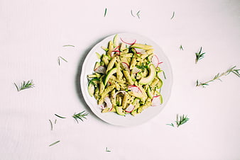 Top view of penne pasta with pesto, avocado, and radishes on white background